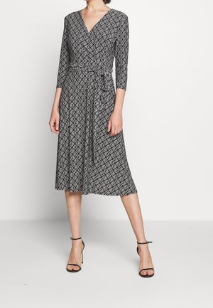 Femme portant une robe portefeuille à motifs géométriques noirs et blancs, manches trois-quarts, et des sandales à talons noirs, debout sur un sol blanc.