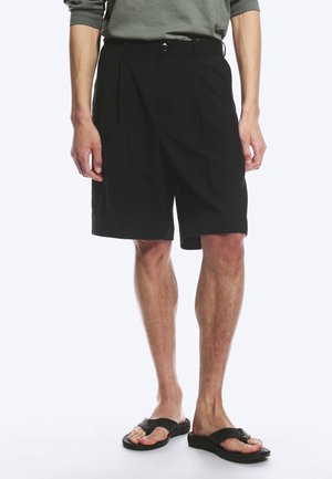Mand iført sorte knælange shorts, grøn langærmet skjorte og sorte sandaler, stående mod en ensfarvet baggrund.