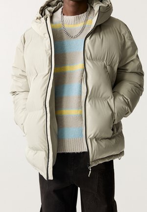 Vue poitrine d'une personne portant une doudoune beige, un pull en tricot rayé bleu, beige, jaune, un collier chaîne argenté et un pantalon noir avec les mains dans les poches.