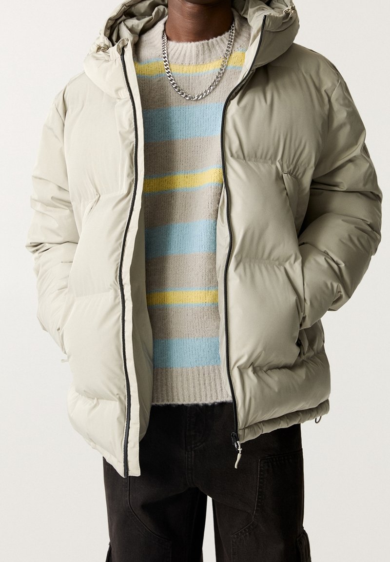Vue poitrine d'une personne portant une doudoune beige, un pull en tricot rayé bleu, beige, jaune, un collier chaîne argenté et un pantalon noir avec les mains dans les poches.