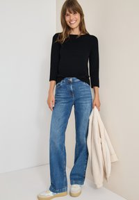 Schwarzes Langarmoberteil kombiniert mit blauen weit geschnittenen Jeans. Eine cremefarbene Fleecejacke hängt über dem Arm. Das Outfit wird mit neutralen Sneakers abgerundet.
