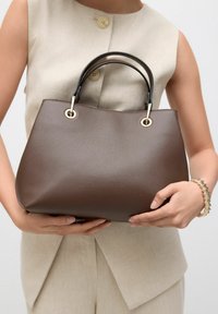Braune Lederhandtasche mit strukturiertem Oberflächen, ausgestattet mit schwarzen Griffen und goldenen Hardware-Details, gehalten von einer Person in einem beigen Outfit.