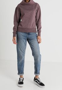 Persona con un suéter de manga larga burdeos, jeans azules remangados y zapatillas blancas y negras de pie sobre un suelo blanco.