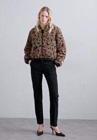 Woman wearing a brown patterned faux fur jacket, black slim pants, and black pointed high heels, standing against a plain gray wall.