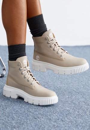 Bottes montantes beiges avec semelles blanches épaisses portées avec des chaussettes noires sur une personne assise sur un tapis bleu.