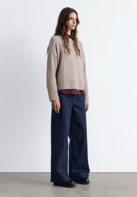 Beige oversized Pullover mit geripptem Muster, kombiniert mit einem dunkelburgunderfarbenen Hemd, dazu weit geschnittene dunkle Jeans und schwarze Schuhe.