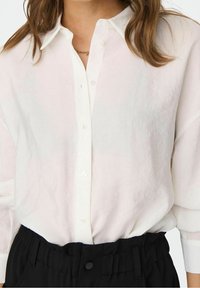 Chemise en lin blanc à boutons avec col classique et coupe décontractée, associée à un pantalon noir taille haute avec ceinture élastique.