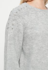 Suéter cinza-claro com malha texturizada, gola redonda e contas prateadas decorativas ao longo da costura do ombro para um detalhe adicional.