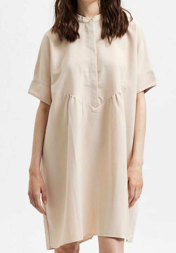 Beige šaty s krátkým rukávem a mandarinovým límcem, které mají plisovaný design. Materiál je hladký s mírným leskem, čímž vytváří minimalistický vzhled.