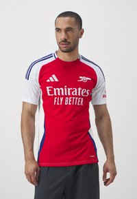 Tricou de fotbal roșu cu accente albe și albastre, având logo-ul "Emirates FLY BETTER" și dungi Adidas pe mâneci. Mâneci scurte, croială athletică.