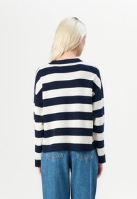 Pulover bleu marin cu dungi crem, din material tricotat, cu guler rotund și croi lejer. Purtați cu blugi din denim cu talie înaltă.