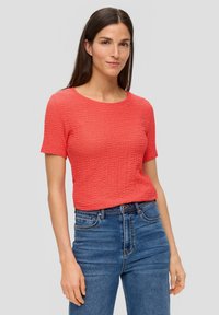 Korallenfarbener, texturierter Kurzarmoberteil mit Rundhalsausschnitt, kombiniert mit hochtaillierten blauen Jeans. Das Oberteil zeigt ein dezentes, zerknittertes Stoffdesign.