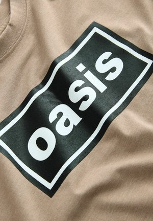 T-shirt en coton beige avec un grand imprimé rectangulaire noir portant le mot "oasis" en blanc, affichant une texture lisse et un design décontracté.