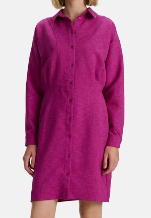 Femme portant une robe-chemise magenta à manches longues boutonnée avec un col, debout devant un fond clair uni.