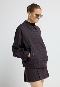 Paars gestreepte jas met een ritssluiting, kraag en twee zijzakken; gecombineerd met een bijpassende mini rok. Model draagt een zonnebril.