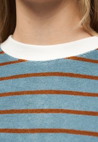 Κοντινό πλάνο στον λαιμό και το επάνω μέρος του στήθους που φορά μπλε πουκάμισο από πετσέτα με καφέ οριζόντιες ρίγες και άσπρο ριμπ ύφασμα στον γιακά.