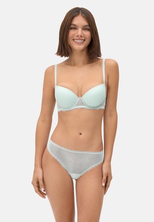 Mujer de pie y sonriendo, con un sujetador azul claro con encaje y bragas a juego transparentes con lunares, sobre un fondo liso.