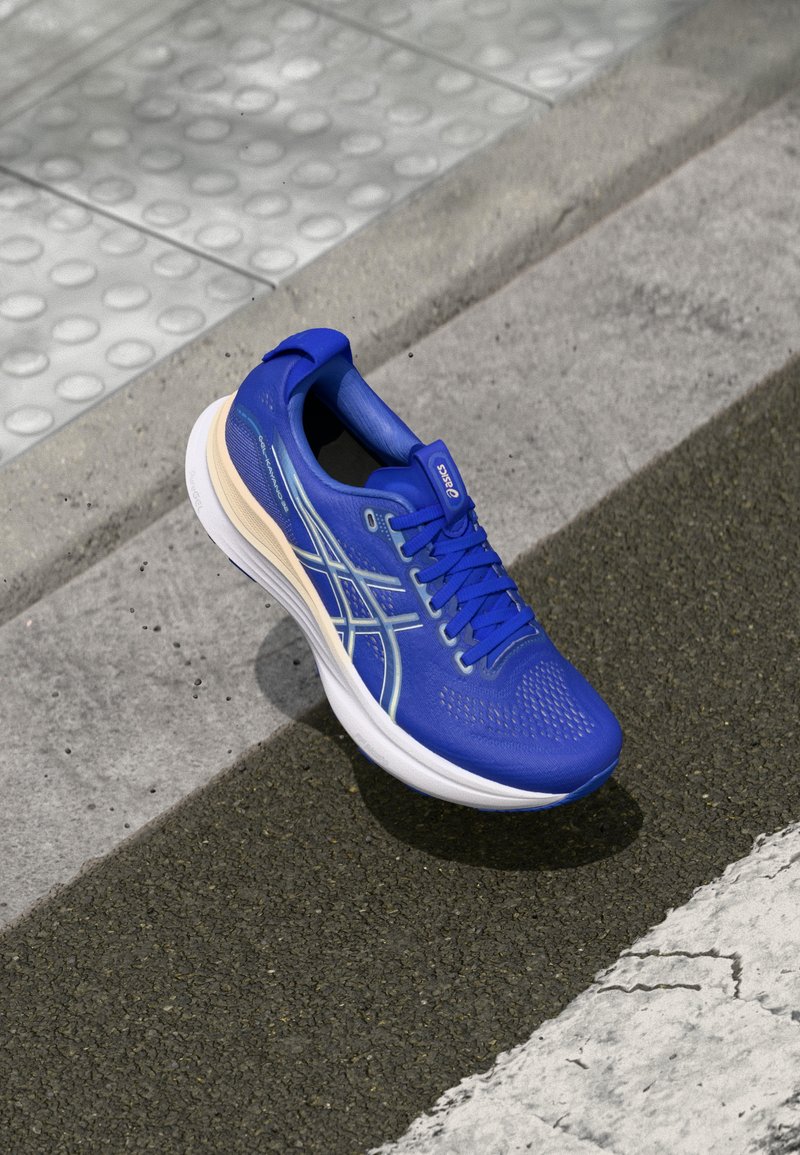 Zapatilla deportiva azul con parte superior de malla, superficie texturizada, entresuela blanca y acentos claros. Presenta cordones y un talón acolchado.