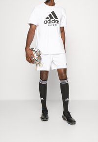 Vit kortärmad skjorta med svart "adidas Fútbol"-logotyp, vita shorts, svarta strumpor med vita ränder, svarta fotbollsskor och en fotboll.