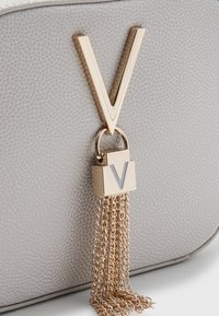 Borsa a tracolla grigia con texture, decorata da una grande lettera 'V' dorata, dotata di lucchetto dorato e dettagli con frange a catena. Design liscio e strutturato.