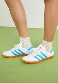 Zapatillas deportivas blancas con rayas azul claro y suelas de color gum, con puntera redonda, cordones y un detalle de marca dorado.