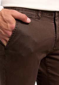 Pantalones de mezcla de algodón marrón con un acabado texturizado, que cuentan con un bolsillo frontal con un dobladillo elegante y un sutil patrón geométrico.