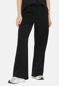 Black sweatpants with a relaxed fit, drawstring waist, and side seam detailing, paired with light-colored athletic shoes.