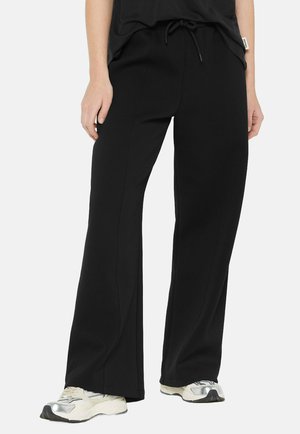 Schwarze Sweatpants mit lockerer Passform, Kordelzugbund und Seitennähten, kombiniert mit hellen Sport Schuhen.