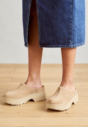 Füße, die beige Wildleder-Slipper mit Plateausohlen tragen, kombiniert mit einem midilangen blauen Jeansrock, stehend auf einem hellen Holzboden.