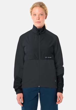 Frau trägt eine schwarze Jacke mit Reißverschluss und Logo auf der Brust sowie blaue Hose, steht frontal vor einem schlichten Hintergrund.