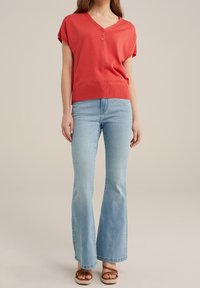 Koronkowa bluzka z krótkim rękawem i dekoltem w serek w kolorze koralowym, z trzema guzikami oraz prążkowanym wykończeniem, zestawiona z jasnoniebieskimi spodniami dzwonami i brązowymi sandałami na paski.