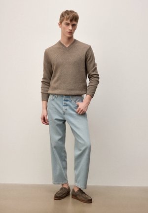 Młody mężczyzna stojący, ubrany w brązowy sweter z dekoltem w serek, jasnobłękitne szerokie dżinsy oraz brązowe mokasyny na jednolitym tle.