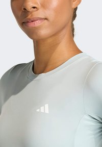 Haut de sport gris clair avec un col rond, des manches courtes, et un logo blanc minimaliste à l'avant. Tissu lisse et extensible.