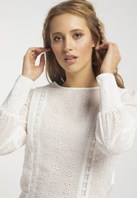 Hvid bluse med hulbroderi, lange pufærmer og rund halsudskæring, med teksturerede detaljer og et delikat blomsterdesign.