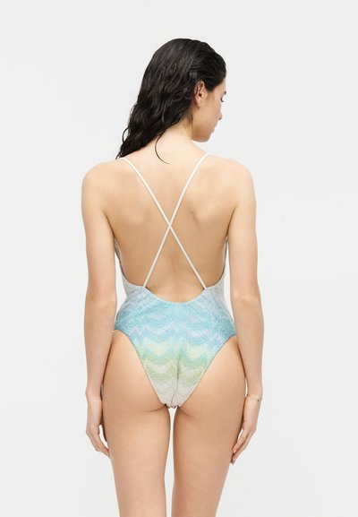 Γυναίκα με βρεγμένα σκούρα μαλλιά που φορά ένα μπλε, πράσινο και ανοιχτόχρωμο παστέλ ολόσωμο μαγιό χωρίς πλάτη με λεπτούς αλληλοδιαπλεκόμενους ιμάντες.