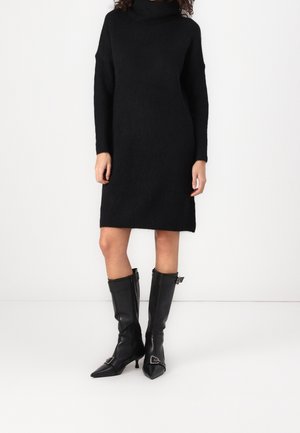 Femme portant une robe-pull noire à manches longues et col roulé, associée à des bottes en cuir noir montantes jusqu’au genou, à bouts pointus et bouclées.