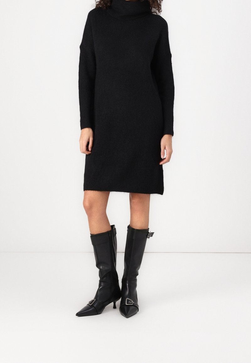 Femme portant une robe-pull noire à manches longues et col roulé, associée à des bottes en cuir noir montantes jusqu’au genou, à bouts pointus et bouclées.