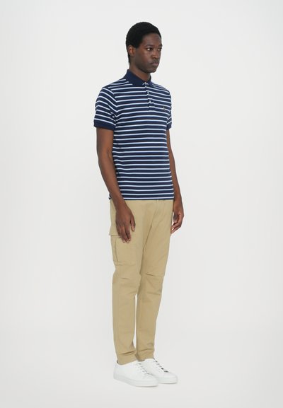 Polo shirt în dungi albastre și bleu deschis, cu guler, asortat cu pantaloni cargo bej și adidași albi. Design simplu, fit casual.