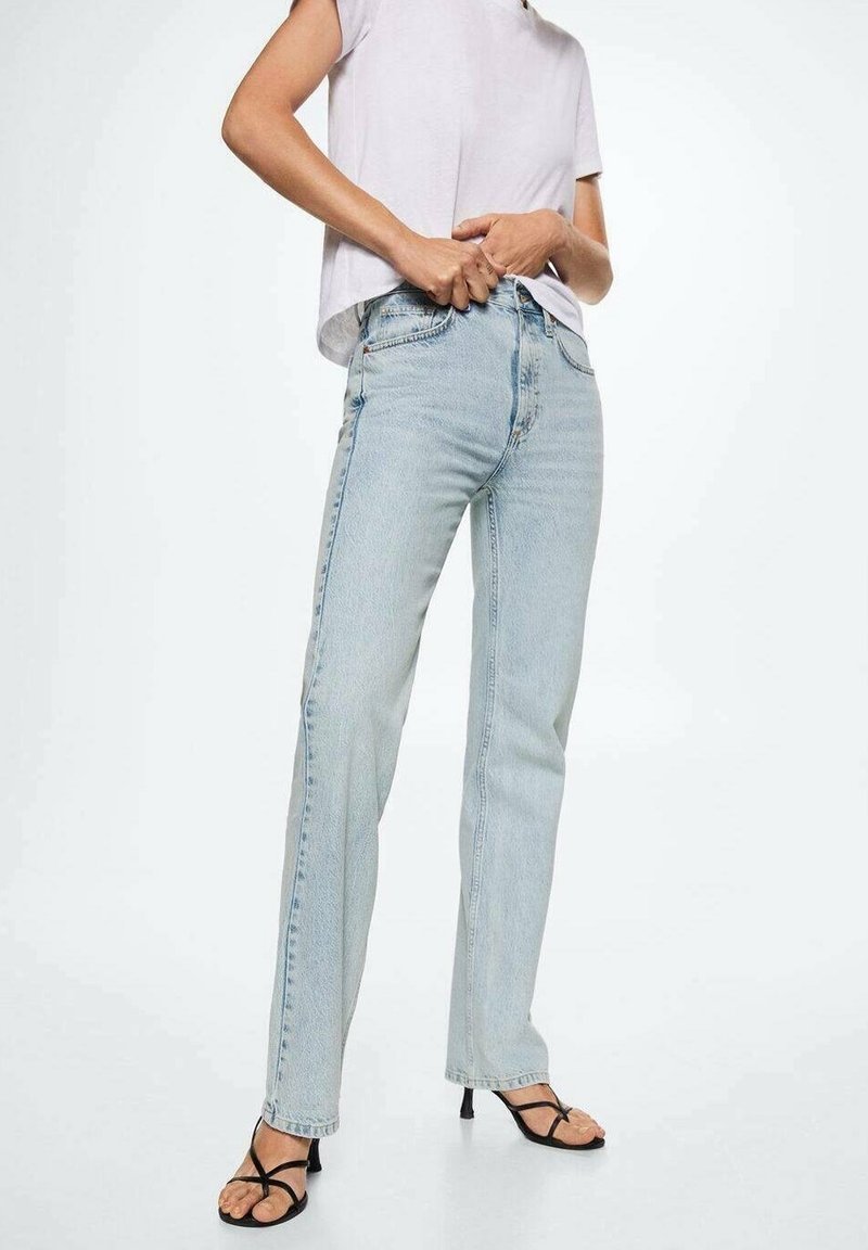 Personne ajustant un jean taille haute bleu clair, portant un t-shirt blanc à manches courtes et des sandales à talons noires à brides, sur fond blanc.