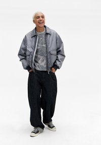 Grå bomberjakke med frontlommer, mønstret grå sweater, brede sorte bukser og sorte sneakers med hvide såler. Model står og smiler.