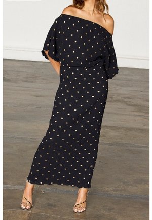 Kobieta ubrana w czarną, długą suknię z odkrytymi ramionami, ozdobioną złotymi grochami, oraz sandały na wysokim obcasie z paskami, stojąca na betonowej podłodze.