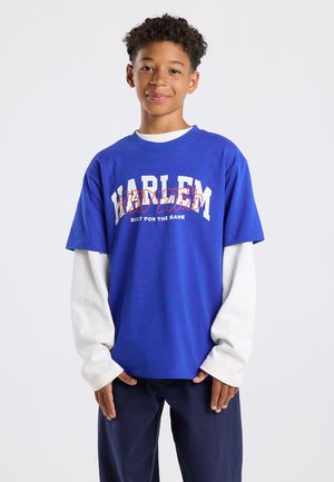 Blauwe t-shirt met "HARLEM" in witte en rode tekst en daaronder "BUILT FOR THE GAME". Gelaagd over een wit longsleeve shirt.