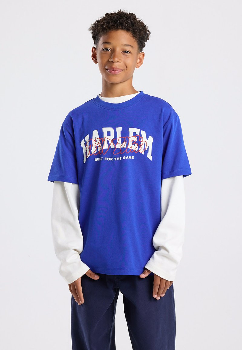 Blue t-shirt featuring "HARLEM" in white and red text with "BUILT FOR THE GAME" underneath. Layered over a white long-sleeve shirt.