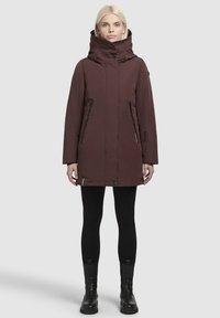 Bordeaux parka met een hoge kraag, voorzien van twee ritszakken aan de zijkant en een cinched taille, gecombineerd met zwarte leggings en enkellaarsjes.