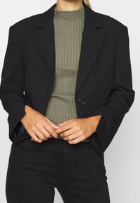 Blazer noir à bouton unique, au design ajusté et avec des revers crantés, porté sur un haut côtelé vert olive.
