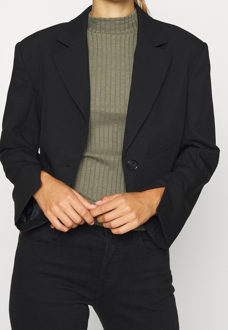 Blazer noir à bouton unique, au design ajusté et avec des revers crantés, porté sur un haut côtelé vert olive.