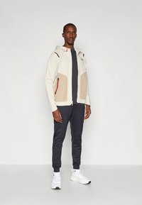 Biała kurtka z beżowymi akcentami, kieszeniami na zamek i regulowanym kapturem. Ciemnoszary top i spodnie, zestawione z białymi trampkami.