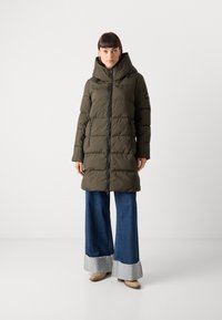 Opgeblazen donkergroen parka met een capuchon, ritssluiting en zijzakken. Heeft een gestikte uitvoering en wordt gedragen over ruime blauwe denim broeken.