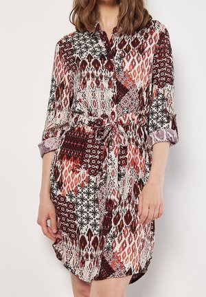 Frau trägt ein knielanges Hemdblusenkleid mit mehrfarbigen geometrischen und floralem Mustern, hochgekrempelten Ärmeln und einem gebundenen Taillengürtel.