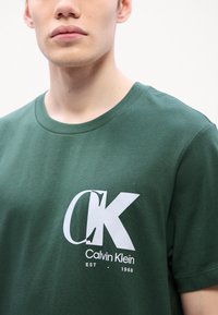 Tamnozeleni pamučni majica s kratkim rukavima s velikim bijelim "CK" logom i tekstom "Calvin Klein EST - 1968" ispisanim na prednjoj strani. Okrugli izrez.